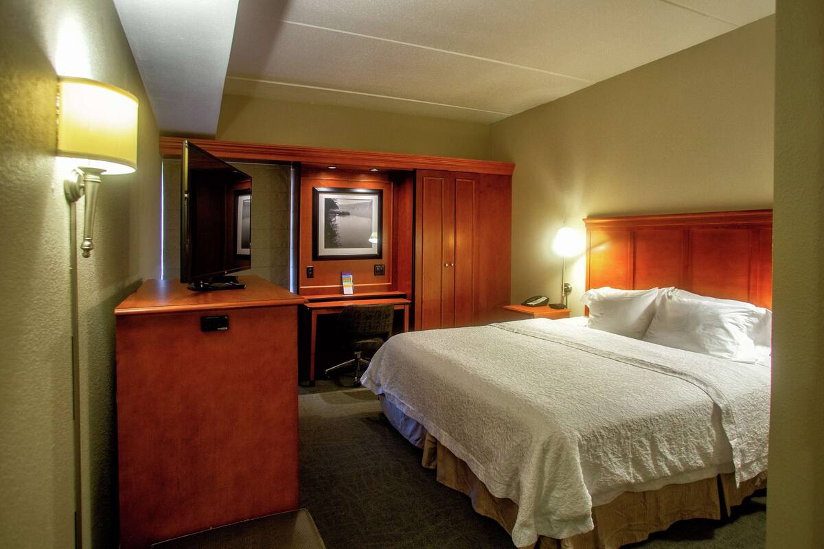 Habitación de hotel con cama, escritorio, silla, lámpara de escritorio y mesita de noche con lámpara.