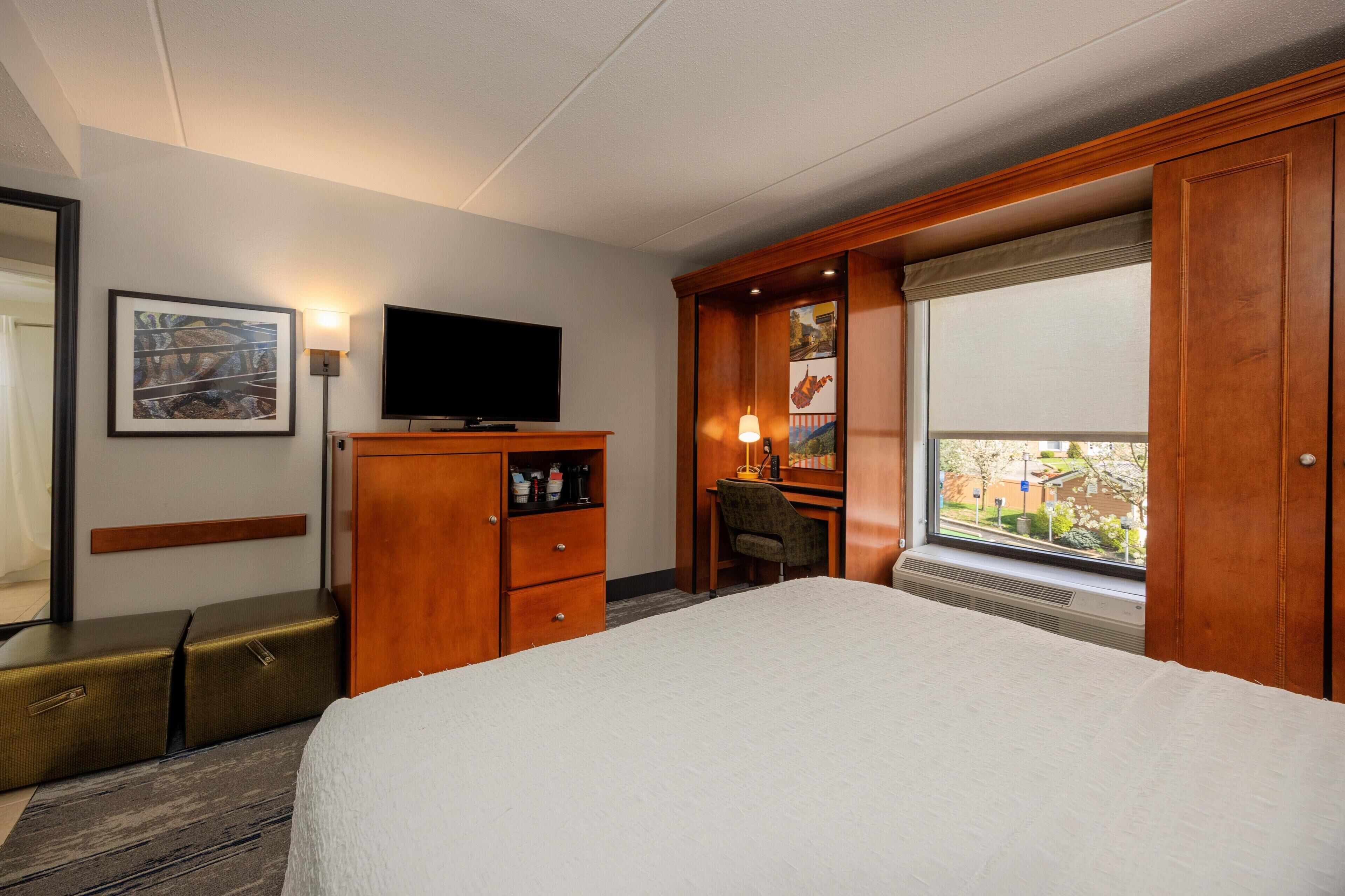Quarto de hotel com cama, escrivaninha, televisão, guarda-roupa e janela com vista.