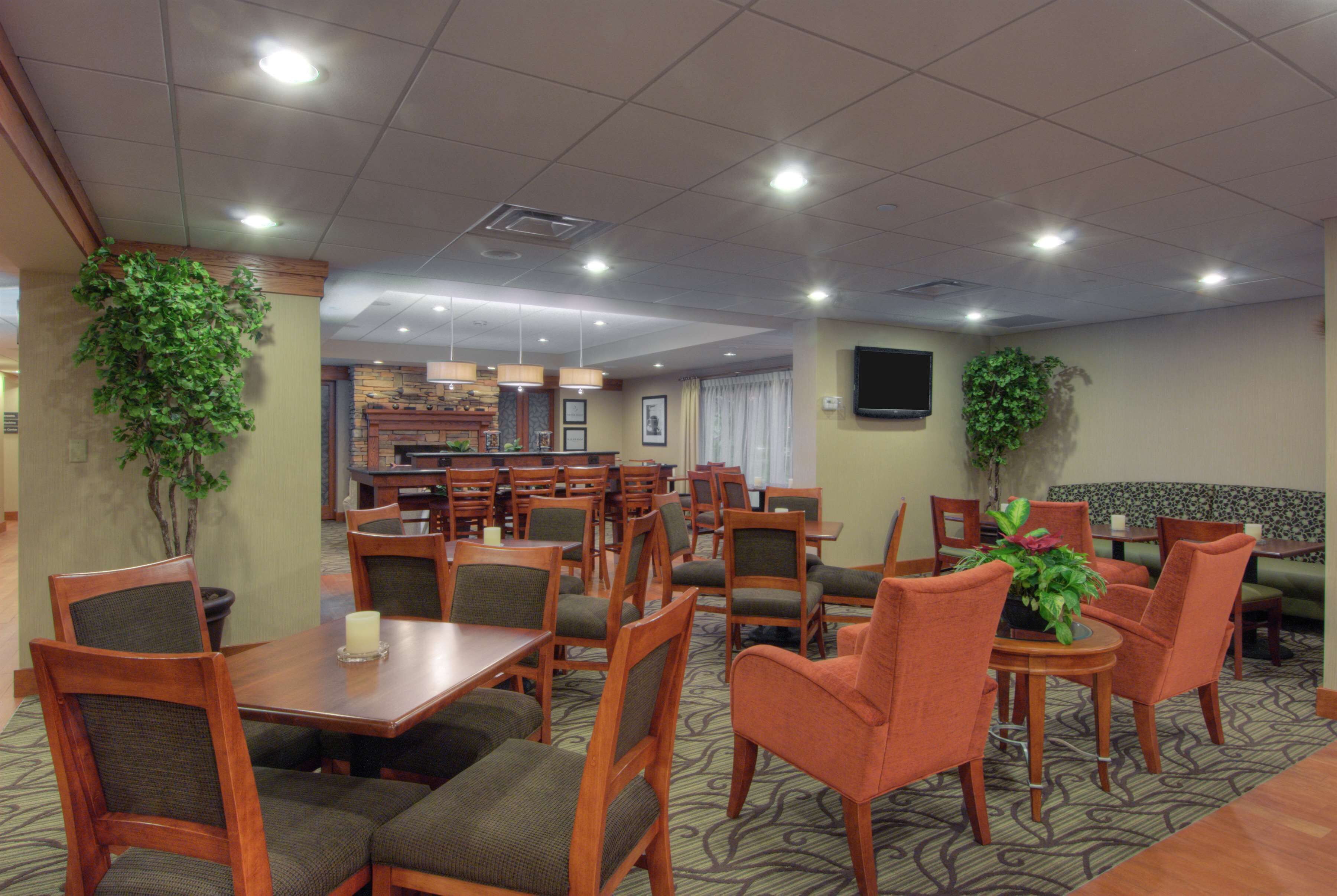 A hotel dining area with multiple tables and chairs, a television mounted on the wall, and potted plants.
