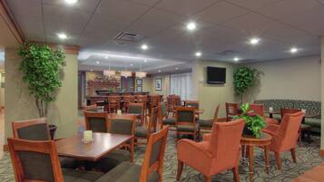 A hotel dining area with multiple tables and chairs, a television mounted on the wall, and potted plants.