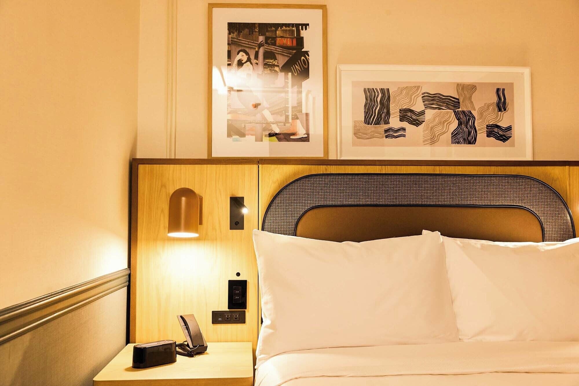 Une chambre d’hôtel avec une tête de lit en bois, une table de chevet avec une lampe, un téléphone et des œuvres d’art encadrées accrochées au mur.