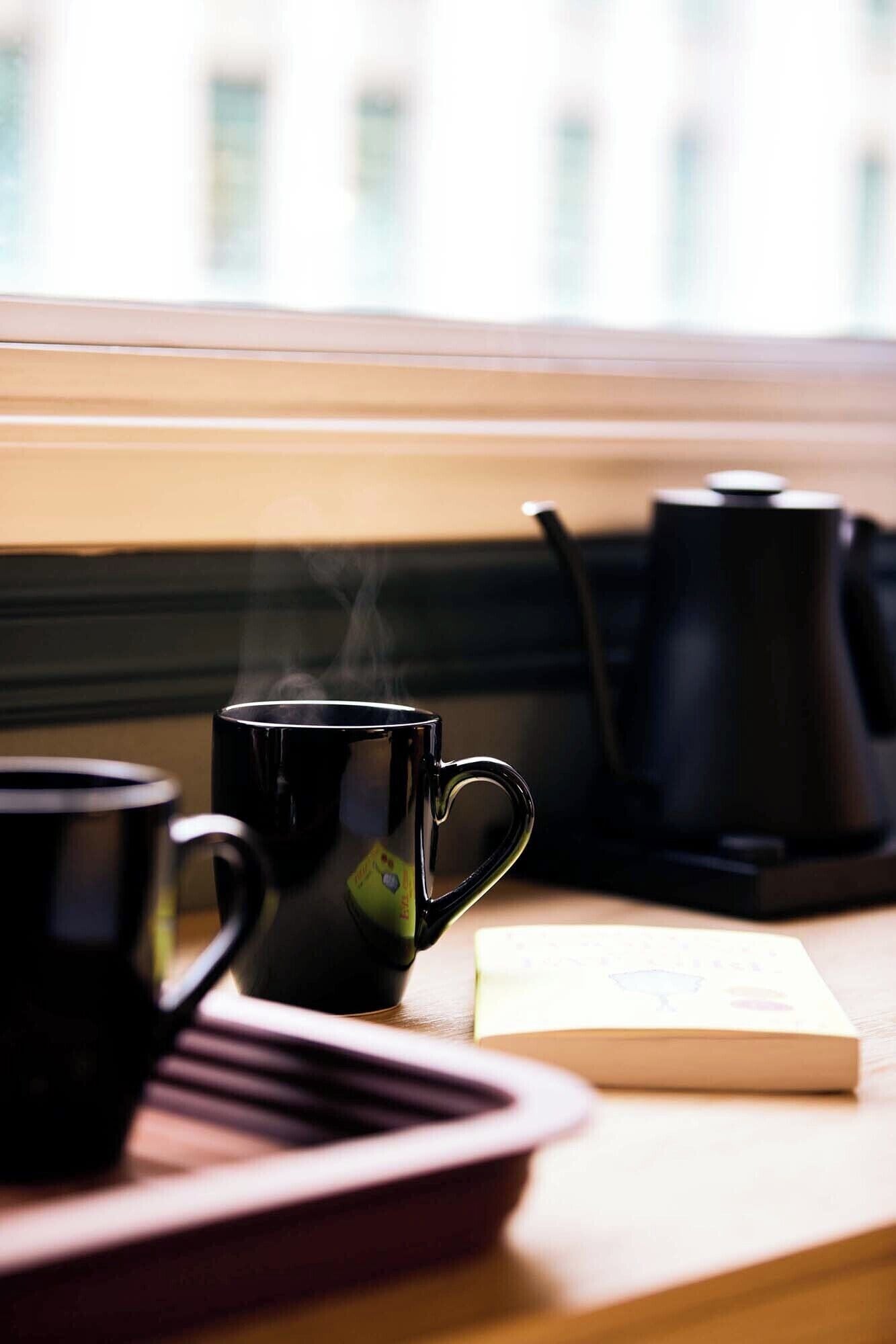Un angolo con caffè: una tazza nera, una bustina di tè e un libro poggiati su un tavolo di legno vicino a una finestra.
