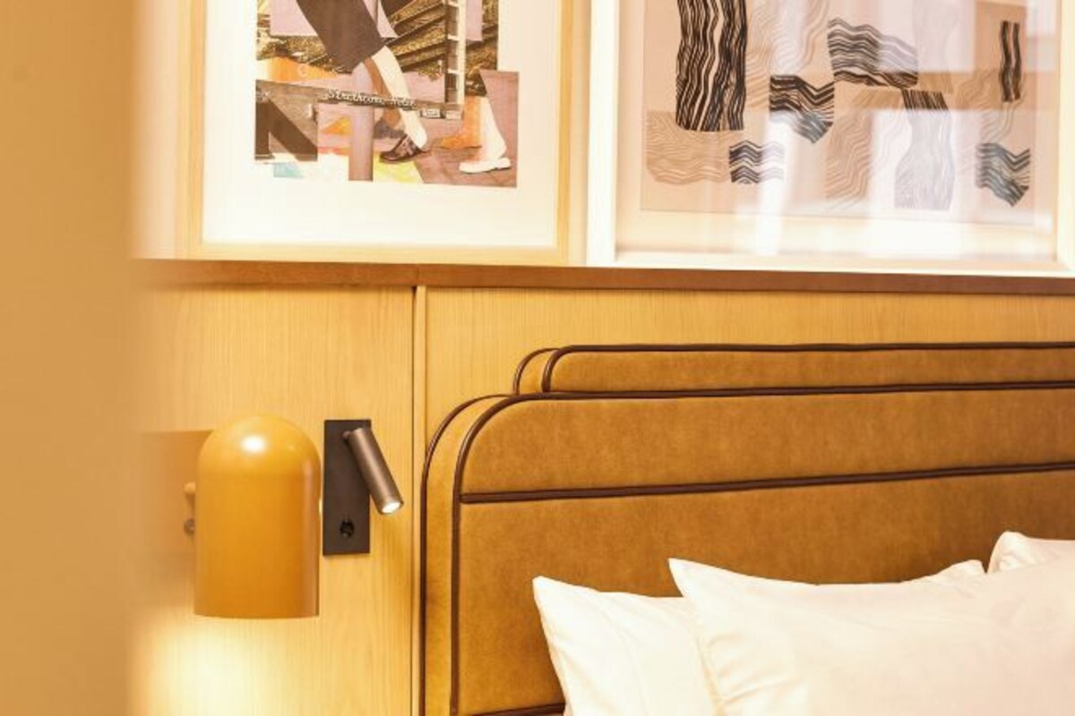A hotel room with a bed, a wall-mounted light, and framed artwork above the bed.