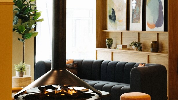 A cozy living room with a modern fireplace, a dark sofa, and a small orange ottoman. There are framed artworks on the wall and a potted plant on the side table. - Union Hotel (Toronto)