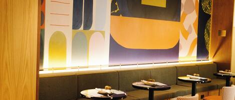 A restaurant with a long counter, cushioned seating, and small round tables.
