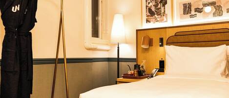 A hotel room with a bed, a nightstand, a lamp, a robe hanging on a rack, and artwork on the wall.