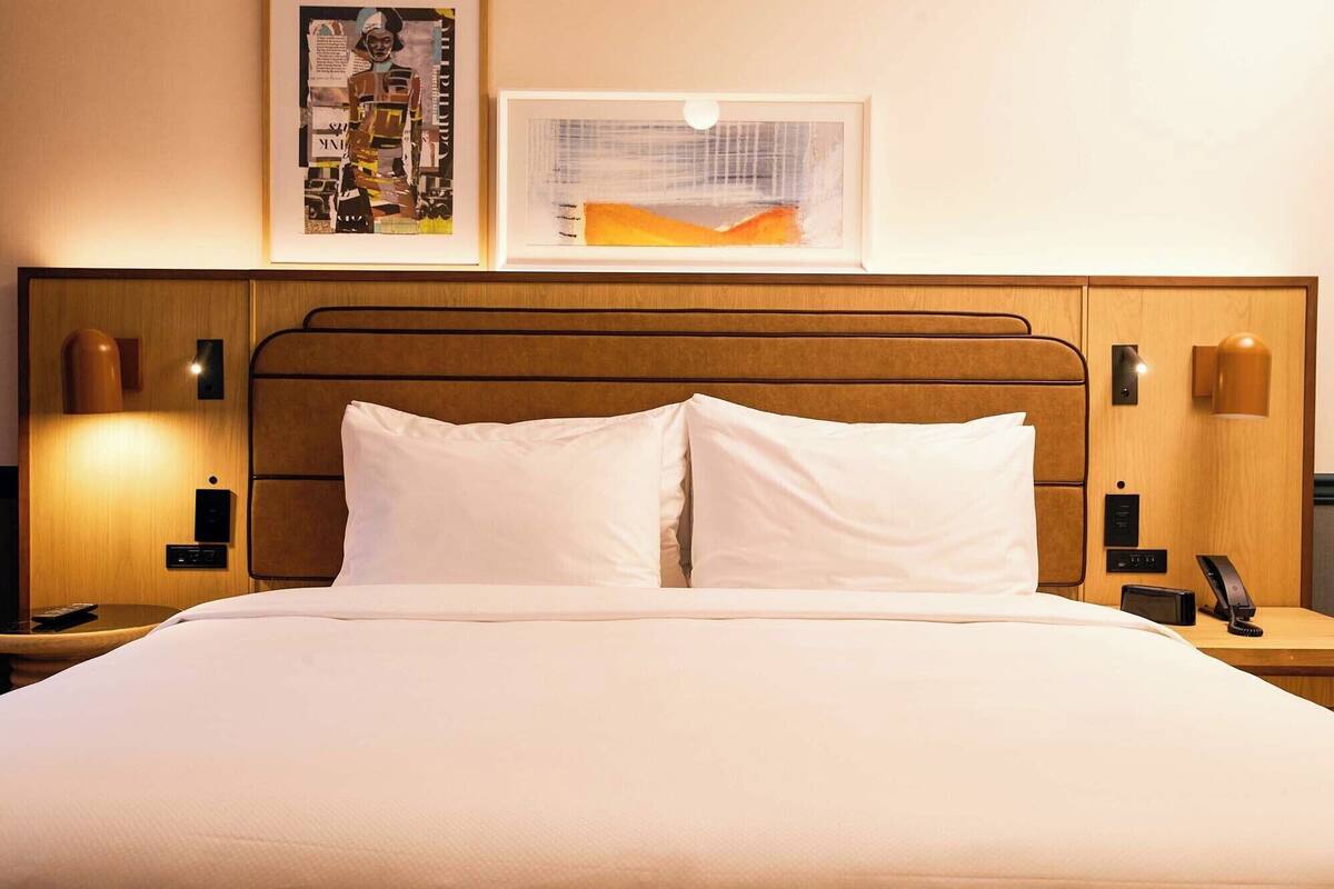 Ein Hotelzimmer mit einem hölzernen Kopfteil, weißer Bettwäsche, zwei Nachttischen mit Lampen und zwei gerahmten Kunstwerken an der Wand.