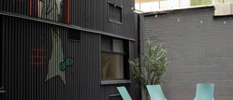 An outdoor seating area with tables and chairs, a building with vertical siding, and a window with a view of another building.