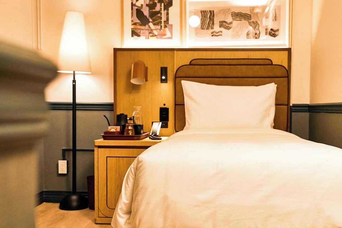 Ein Hotelzimmer mit einem ordentlich bezogenem Bett, einem Nachttisch mit Lampe, Telefon und einer Kaffeemaschine. An der Wand über dem Bett befinden sich gerahmte Bilder.
