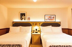Una habitación de hotel con dos camas, cabecero de madera, mesitas de noche con lámparas y cuadros en la pared.