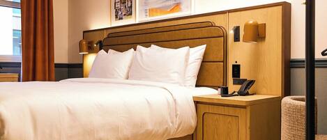 A hotel room with a wooden bed, white bedding, a bedside table with a lamp, a phone, and a waste bin.