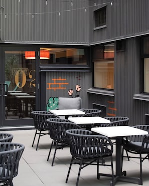 An outdoor seating area with tables and chairs, a glass door entrance, and a building facade with vertical lines and a window.