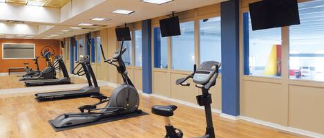 A gym with multiple stationary bikes and treadmills, large windows, and a wooden floor.