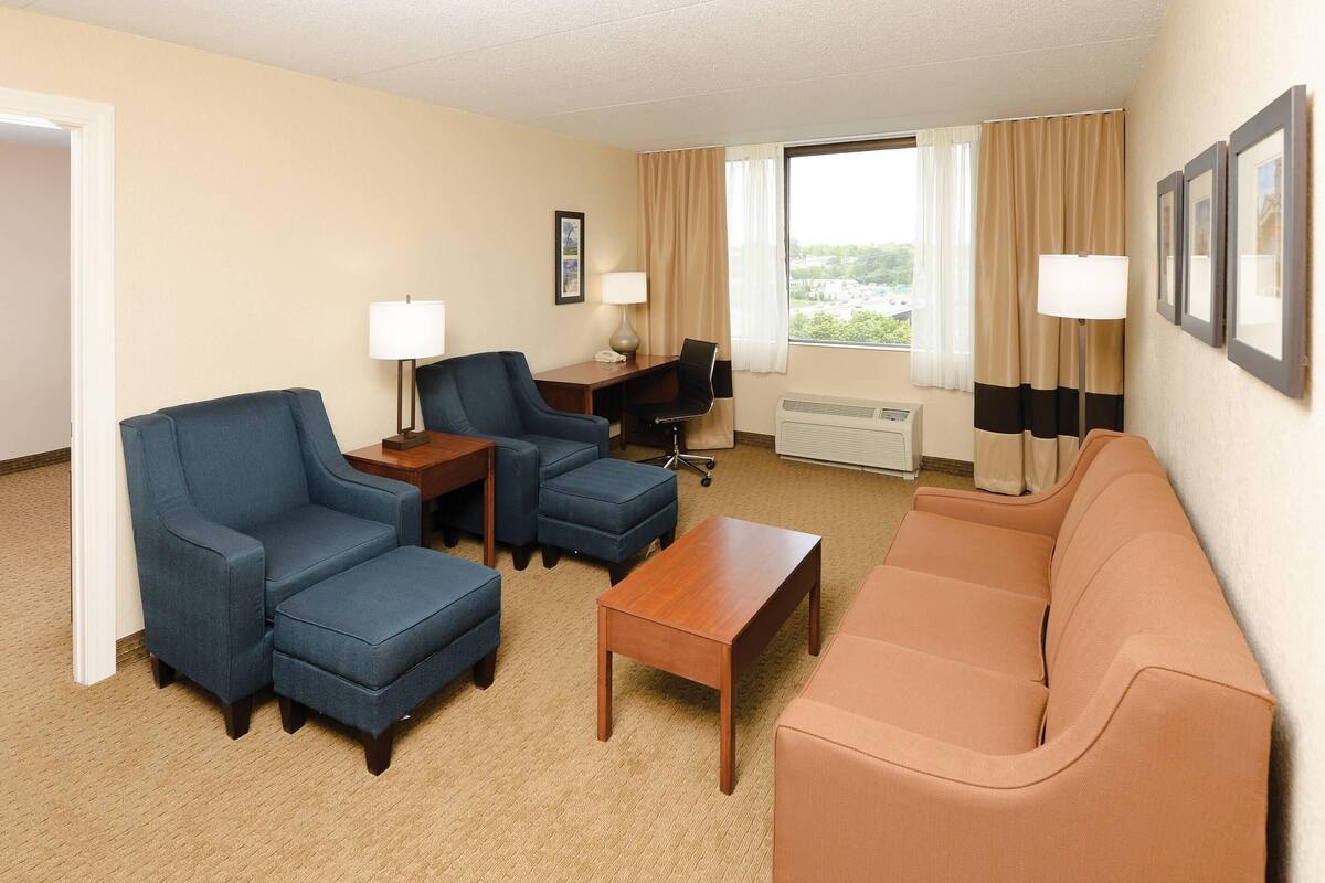A hotel room with a blue armchair, a brown sofa, a wooden coffee table, a desk with a computer, and a window with curtains.
