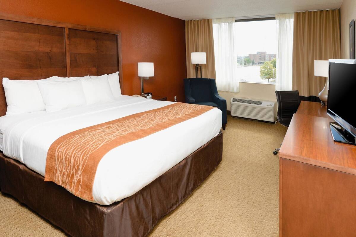 A hotel room with a large bed, a desk with a TV, a chair, a lamp, and an air conditioning unit.