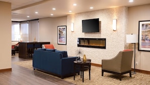 Un hall de hotel con un sofá azul, un sillón color beige, un televisor de pantalla plana montado en una pared con textura, una chimenea y una zona de comedor al fondo.