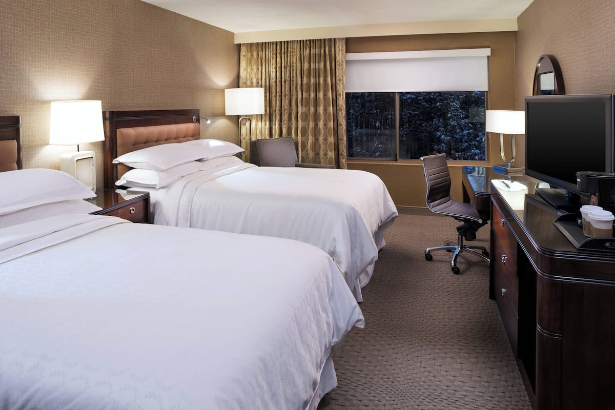 A hotel room with two beds, a desk, a TV, and a window with curtains.