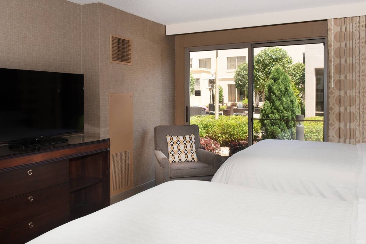 A hotel room with a large bed, a TV on a dresser, a chair with a patterned pillow, and a view of the outside.
