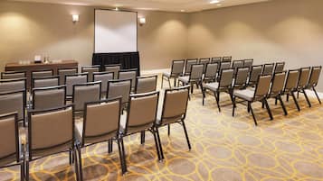 Un salón de conferencias con filas de sillas, una pantalla de proyector y una mesa con vasos de agua.