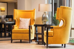 Un sillón amarillo con un cojín, una mesita con una lámpara y vistas al exterior a través de una ventana.