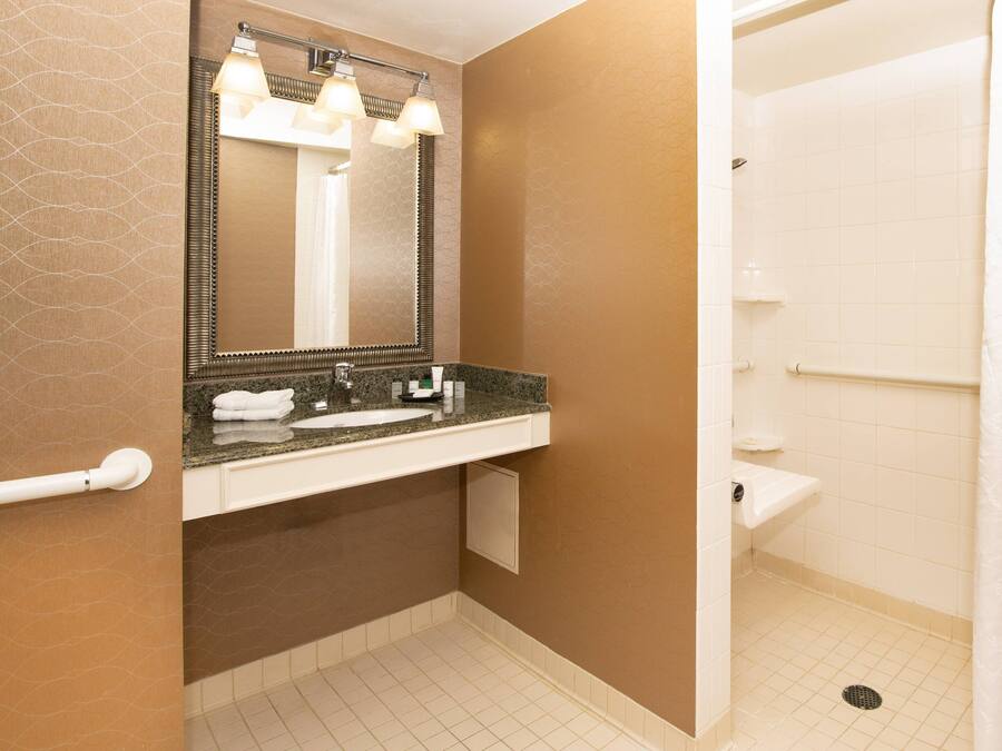 A bathroom with a granite countertop sink, a mirror with lights, a towel rack, and a shower area.