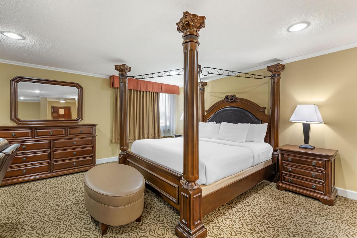 A four-poster bed with a wooden frame, a bedside table with a lamp, a dresser, and a round ottoman.
