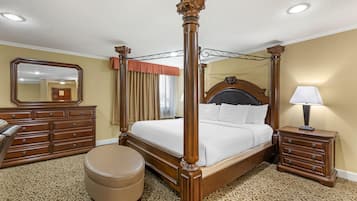 A four-poster bed with a wooden frame, a bedside table with a lamp, a dresser, and a round ottoman.
