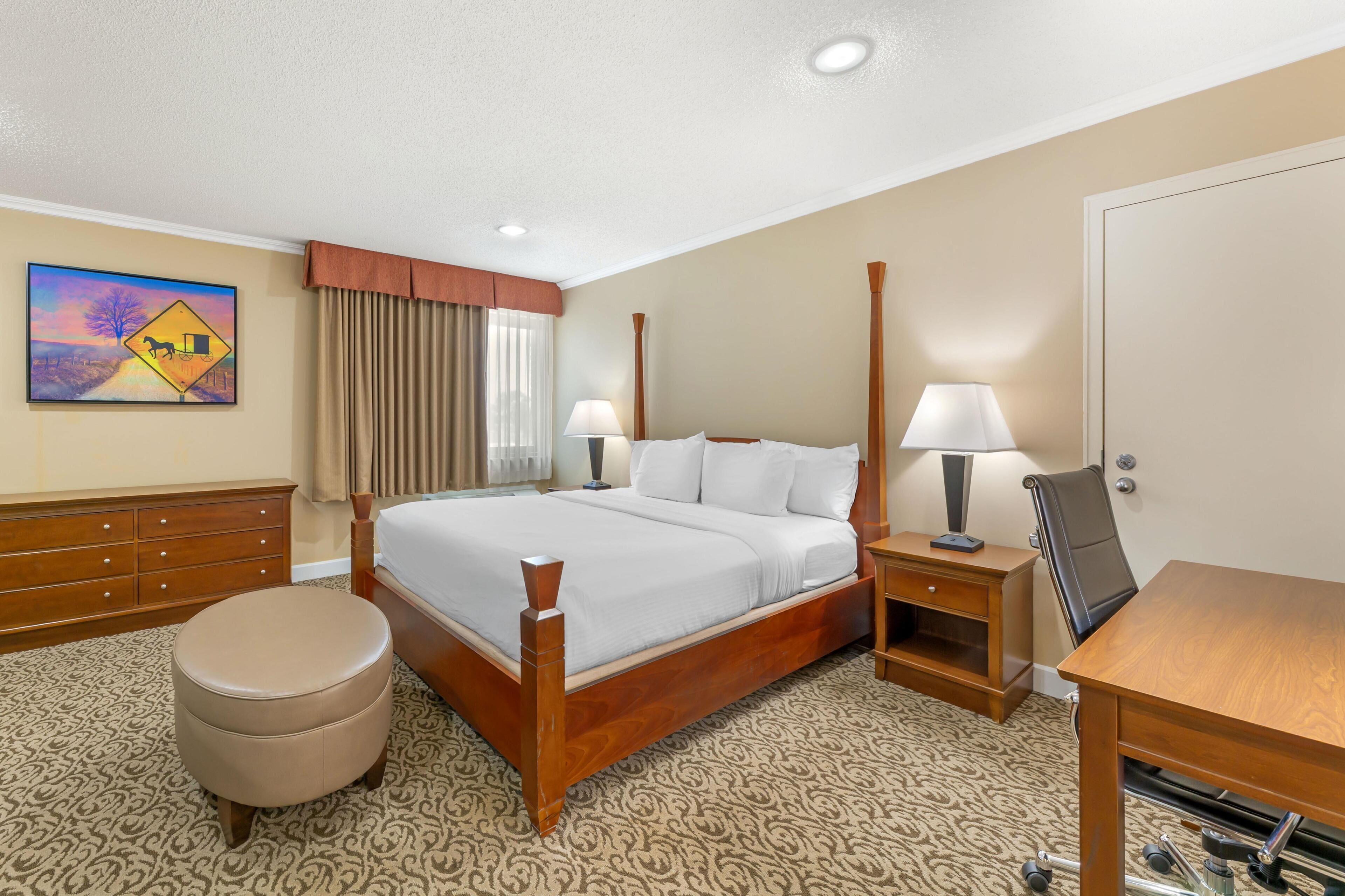 A hotel room with a large bed, two bedside tables with lamps, a wooden dresser, a round ottoman, and a painting on the wall.