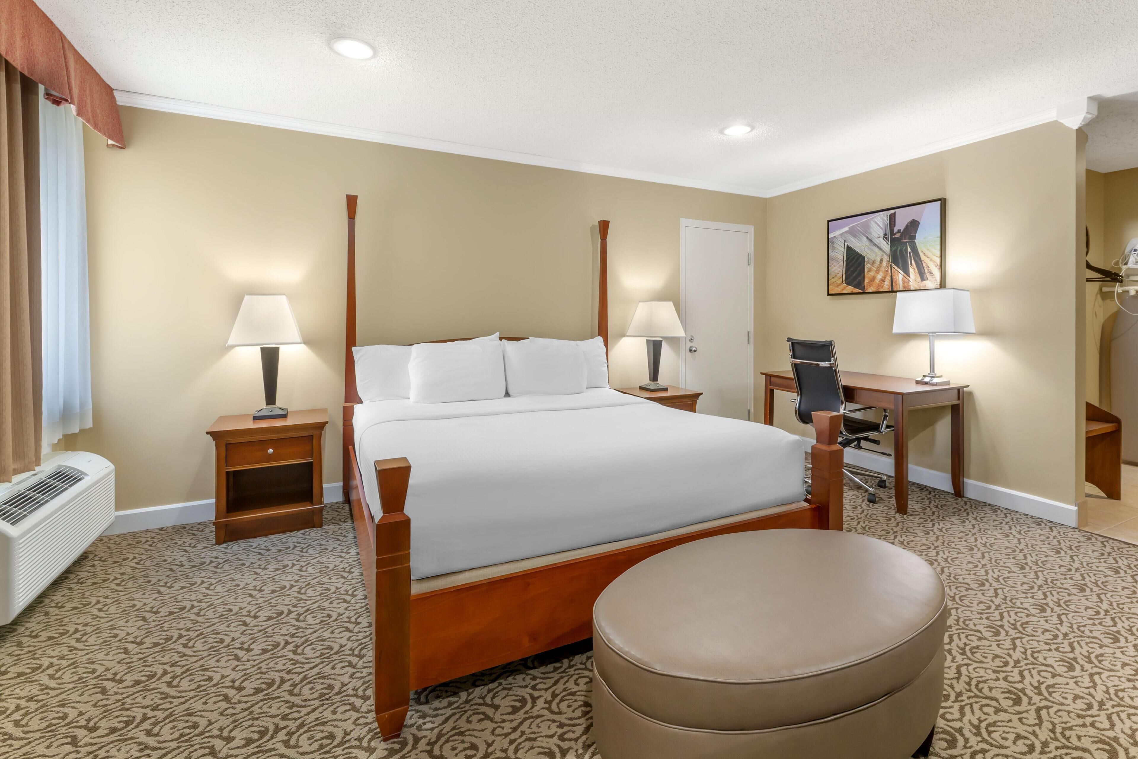 A hotel room with a four-poster bed, a desk with a chair, a standing lamp, and a painting on the wall.