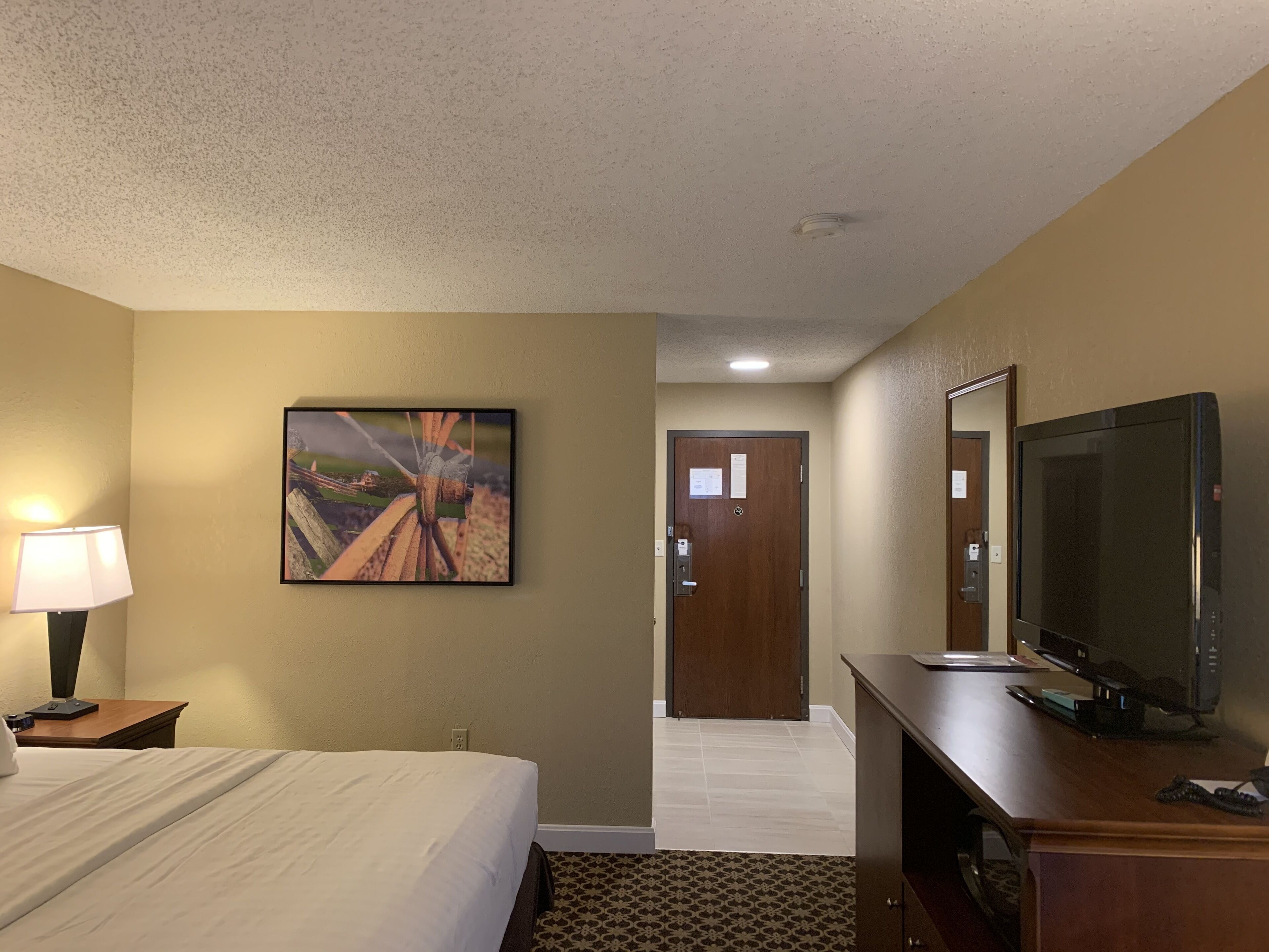 A hotel room with a bed, a desk with a computer, a TV, a lamp, and a painting on the wall.