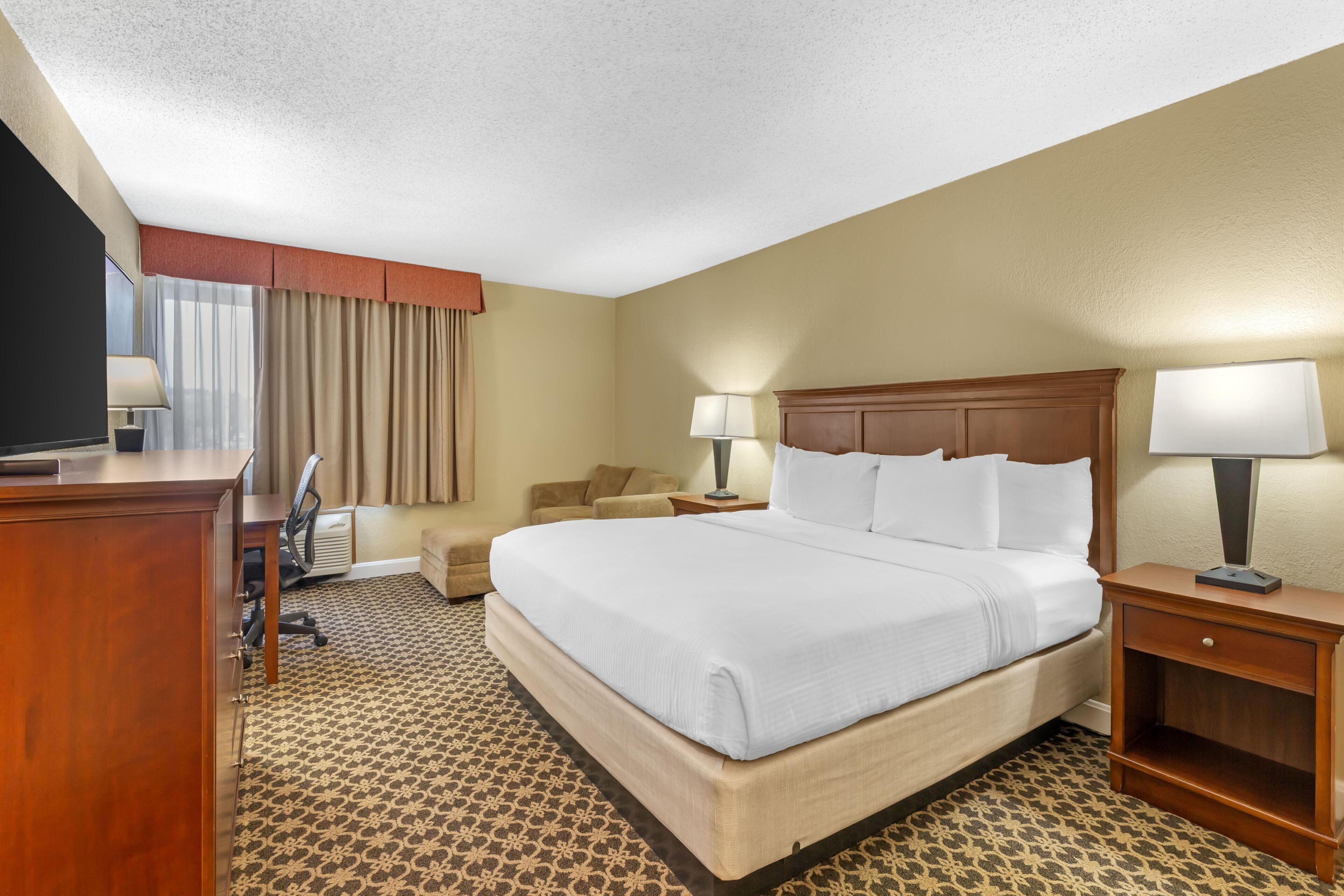 A hotel room with a large bed, a desk, a chair, a TV, and a lamp.