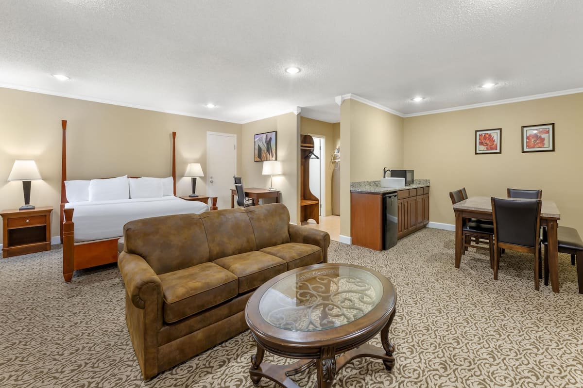 A hotel room with a bed, a sofa, a glass-top coffee table, a dining table with chairs, a kitchen area, and a bathroom visible through an open door.