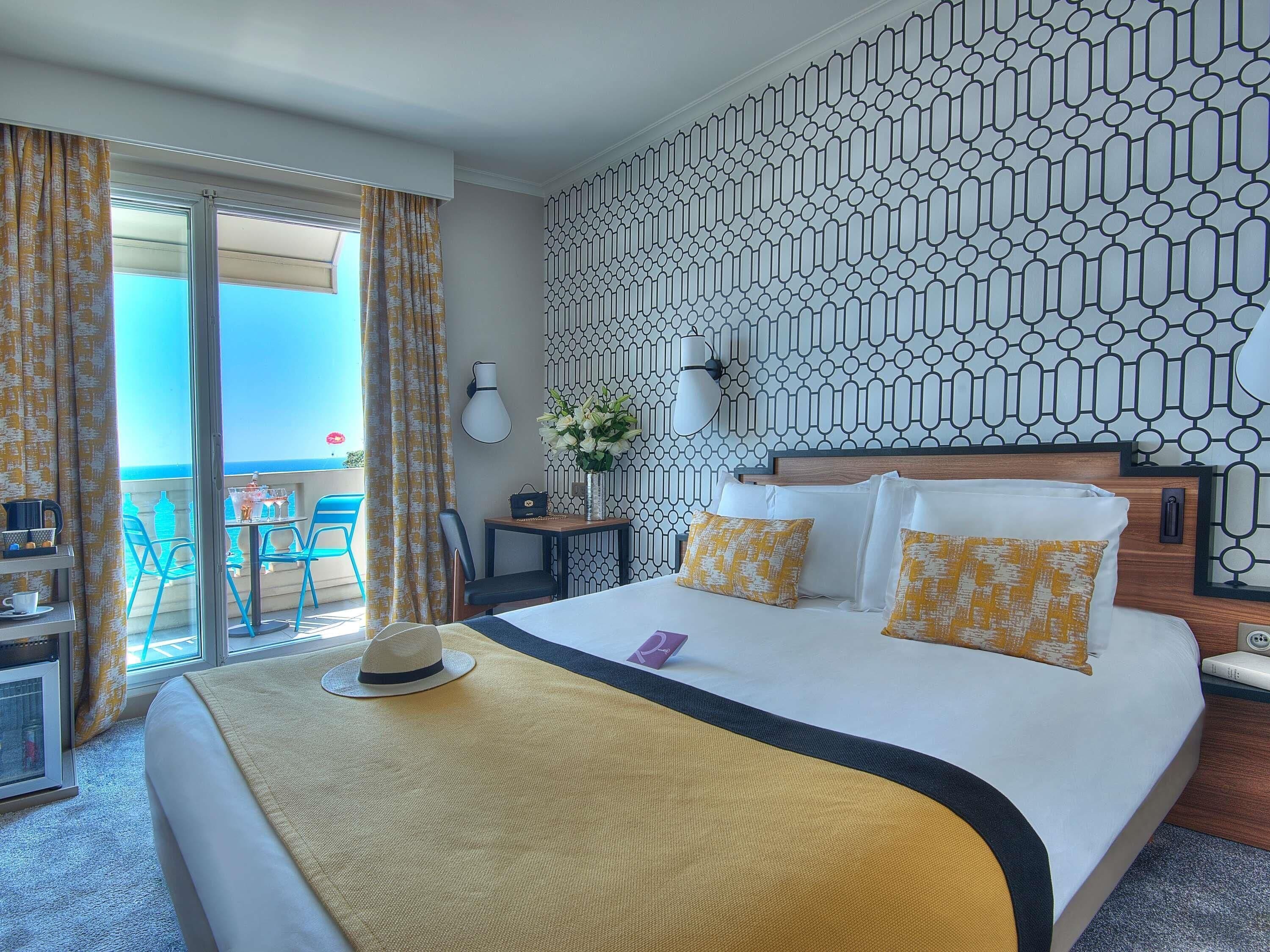 A hotel room with a bed, a desk, a chair, a table with a chair, a window with curtains, and a patterned wall.