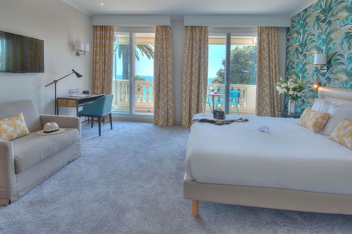 Quarto de hotel com cama, escrivaninha, poltrona, sofá e varanda com vista.