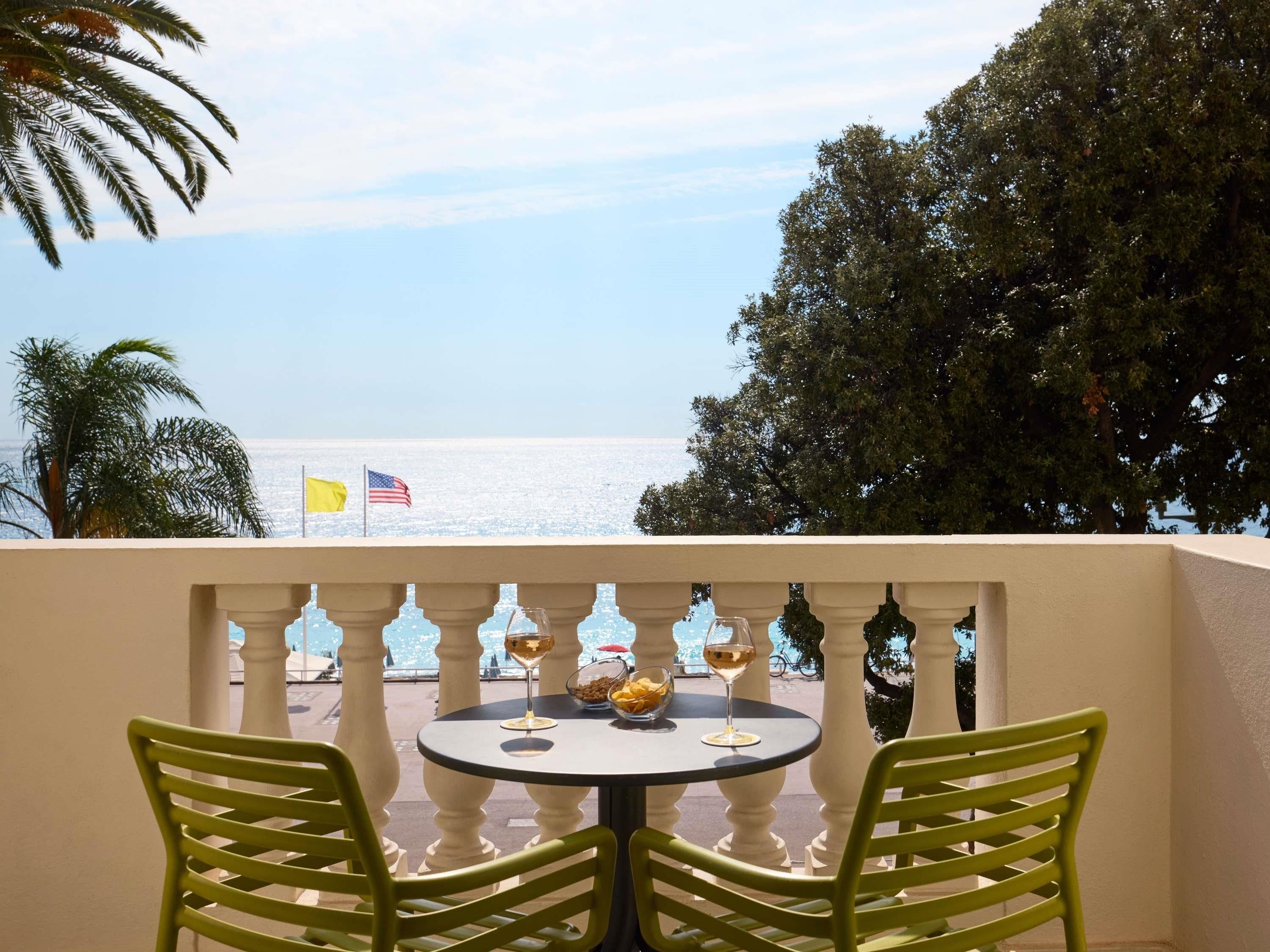 Un balcón con dos sillas y una mesa, con vista a una playa con banderas.