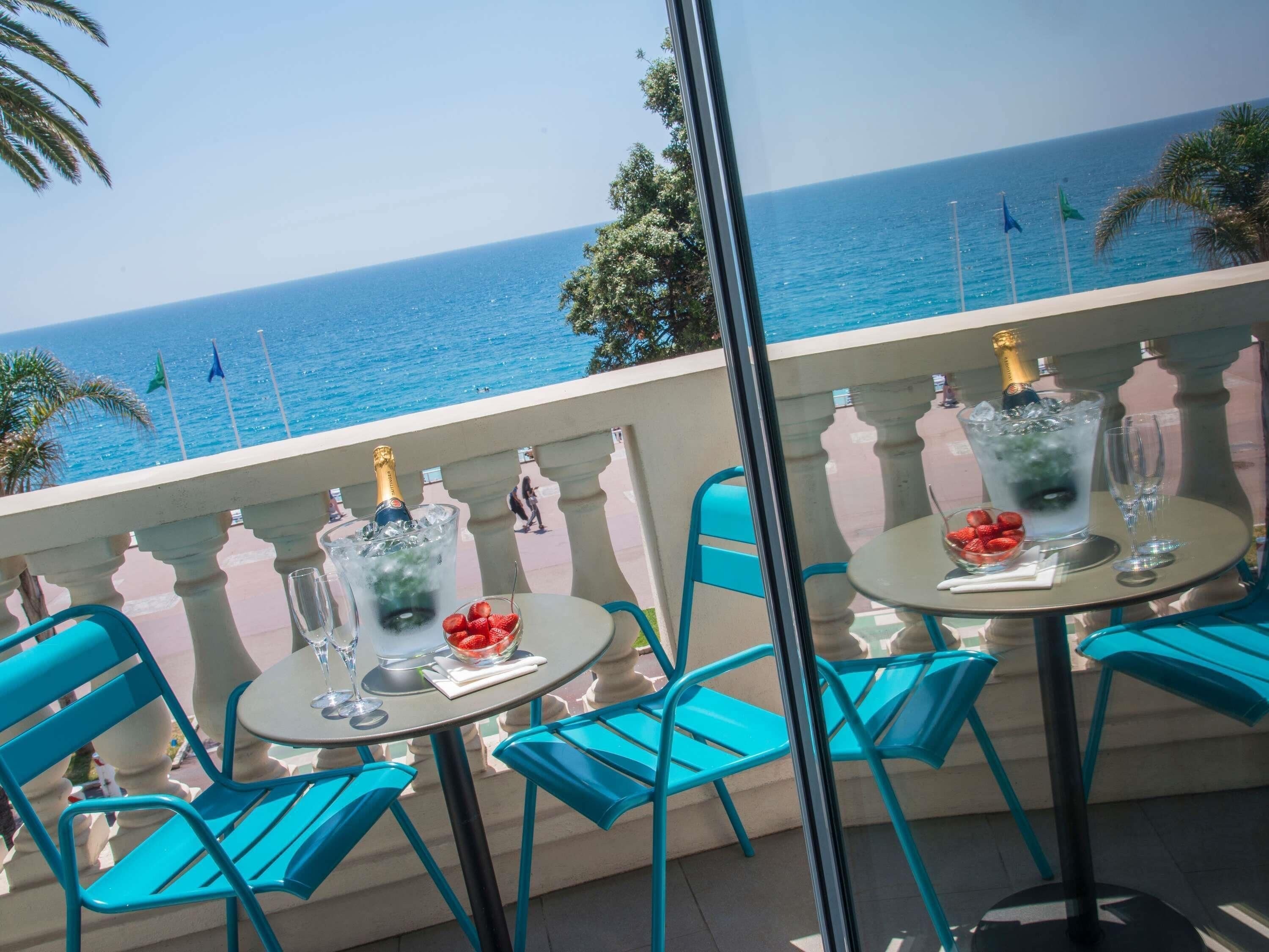 A balcony with a table set for two, overlooking a beach with the sea and flags.