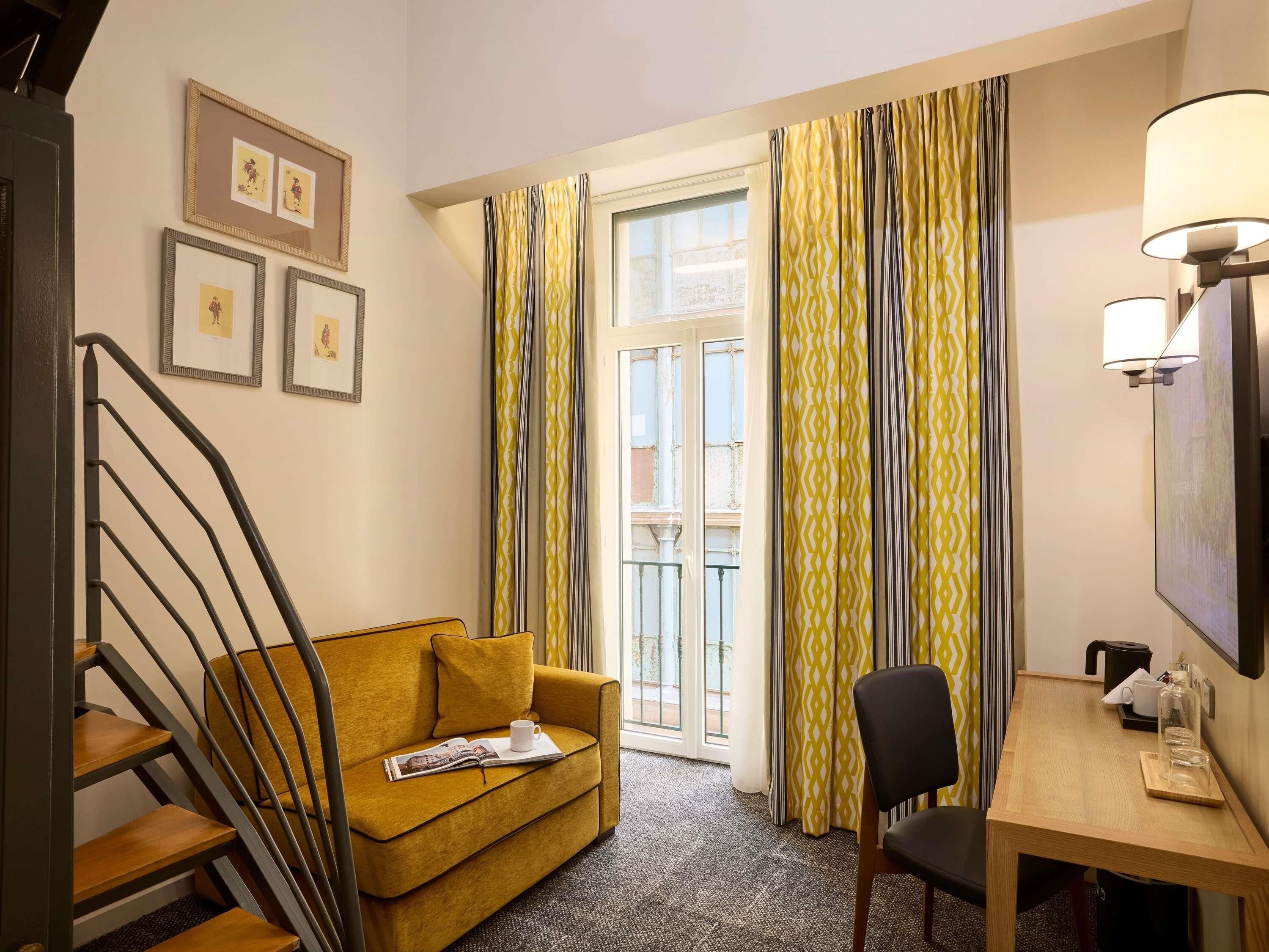 A hotel room with a sofa, a dining table, a chair, a staircase, and a window with curtains.