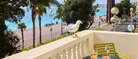 A balcony with a table and chairs, overlooking a beach and promenade with people and a seagull.