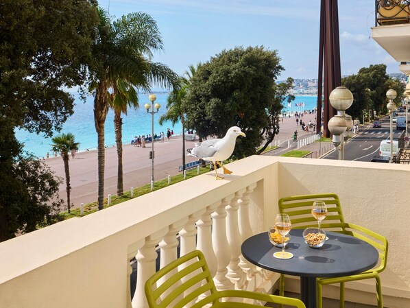 A balcony with a table and chairs, overlooking a beach and promenade with people and a seagull. - Mercure Nice Marche aux Fleurs (Nice)