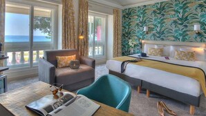 A hotel room with a bed, a sofa, a chair, a table with a magazine and glasses, and a view of the sea through large windows. - Mercure Nice Marche aux Fleurs (Nice)