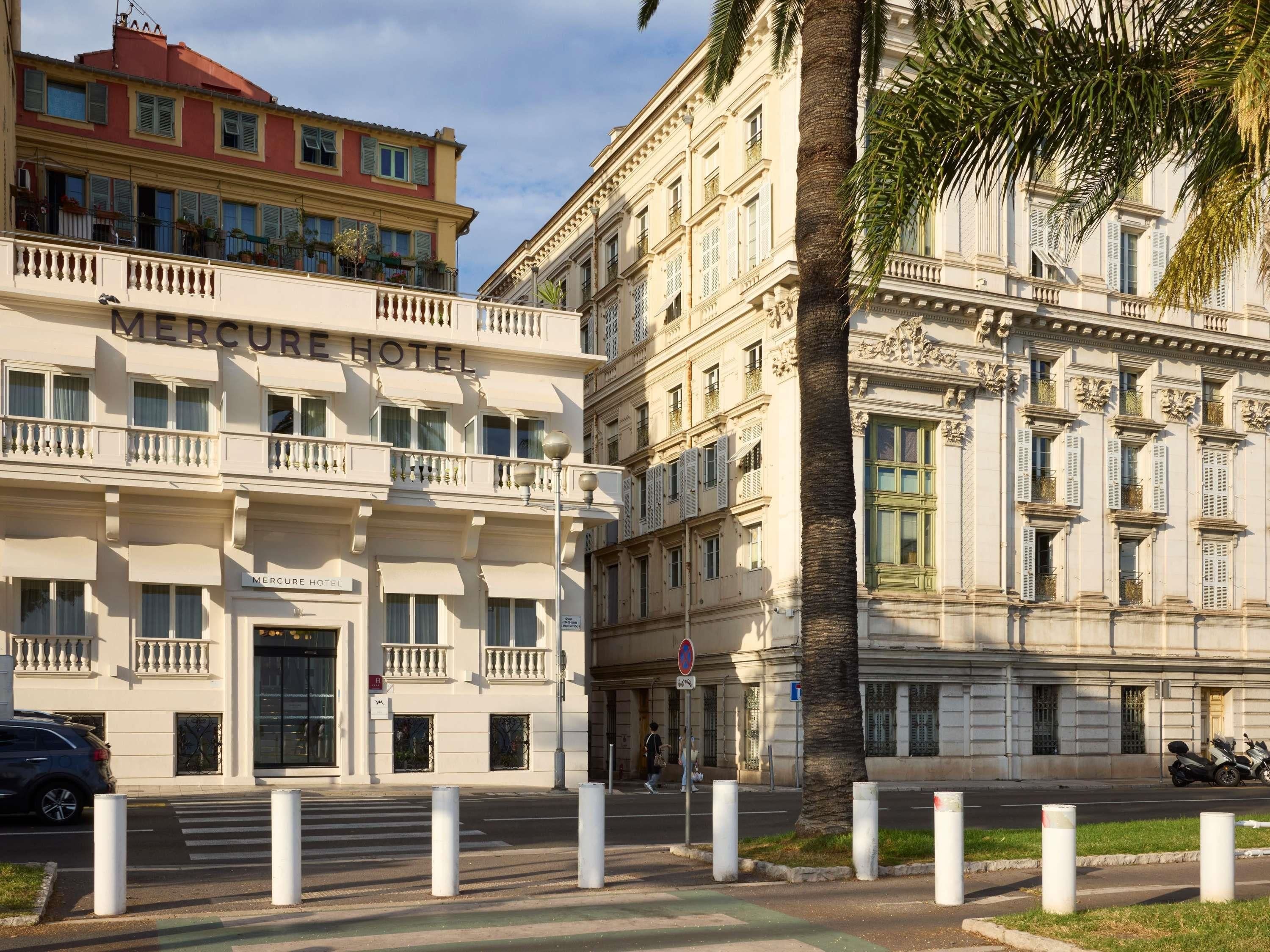 Un hotel con fachada clásica, varios balcones y vista a la calle con separadores y una palmera.
