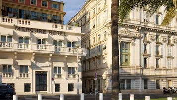 A hotel with a classic facade, multiple balconies, and a street view with bollards and a palm tree.