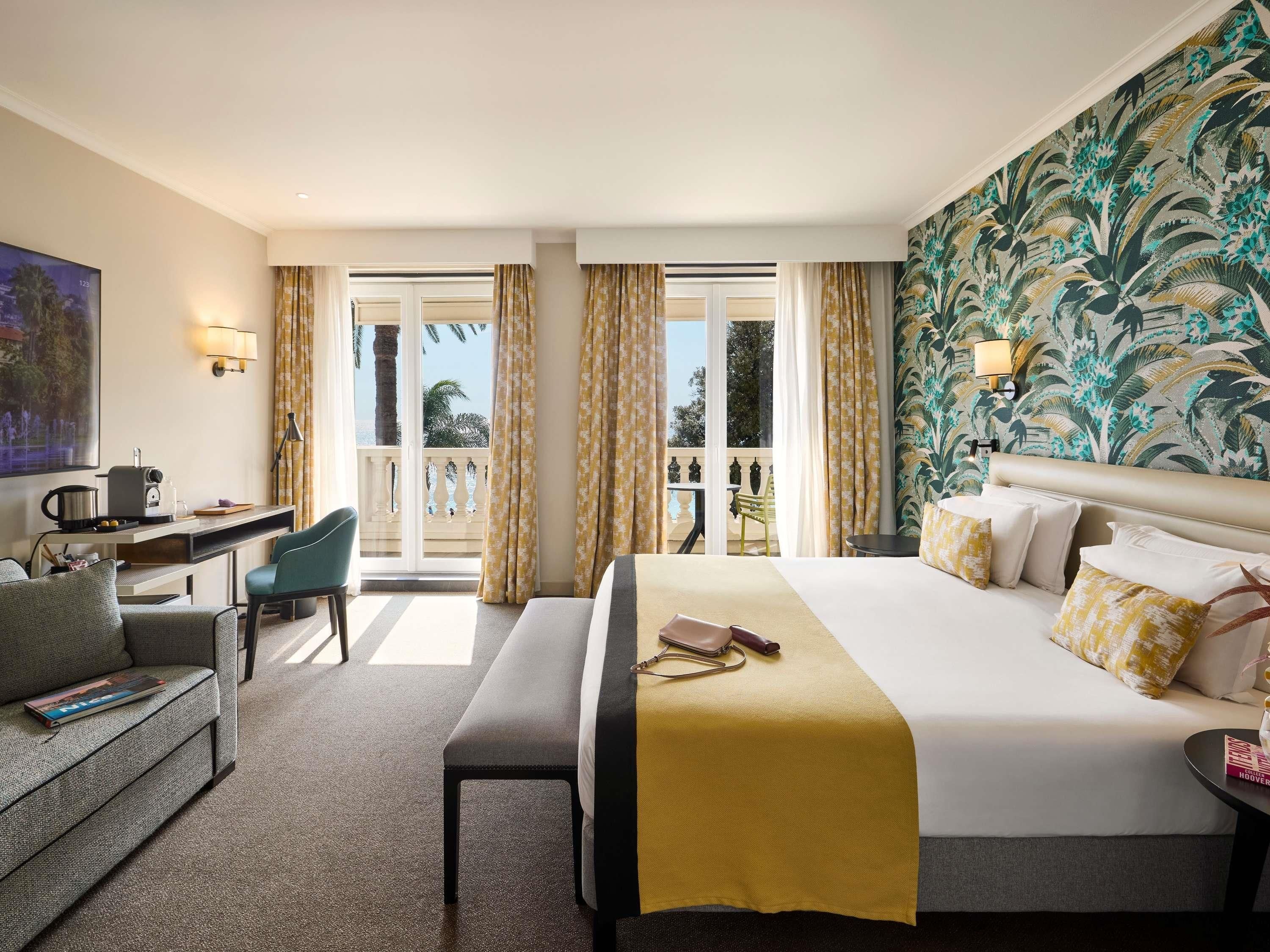 Habitación de hotel con cama, escritorio, silla, mesita y balcón con cortinas.