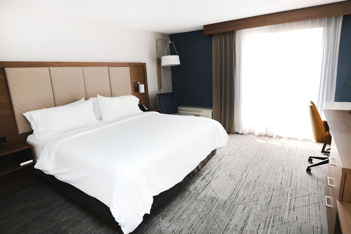 A hotel room with a large bed, a desk with a chair, a lamp, and a window with curtains.