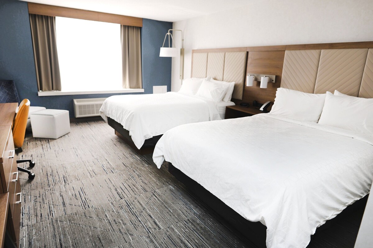 A hotel room with two beds, a desk, a chair, a lamp, and a window with curtains.