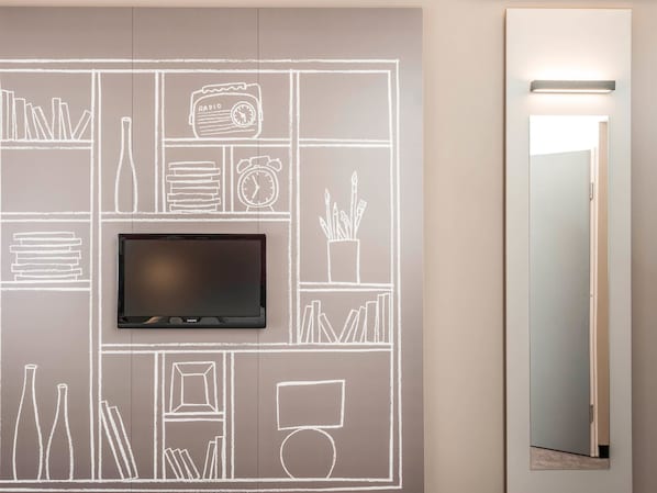 Une chambre moderne avec un téléviseur à écran plat fixé sur un mur recouvert d’un tableau noir, une porte dotée d’un miroir et un luminaire placé au-dessus.