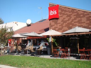 An outdoor seating area with umbrellas and chairs, a red 'ibis' sign, and a brick building facade. - ibis Bourg En Bresse (Bourg-en-Bresse)