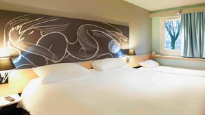 Ein großes Bett mit weißer Bettwäsche, ein an der Wand befestigtes Kunstwerk, ein Nachttisch mit Lampe und ein Fenster mit Vorhängen.