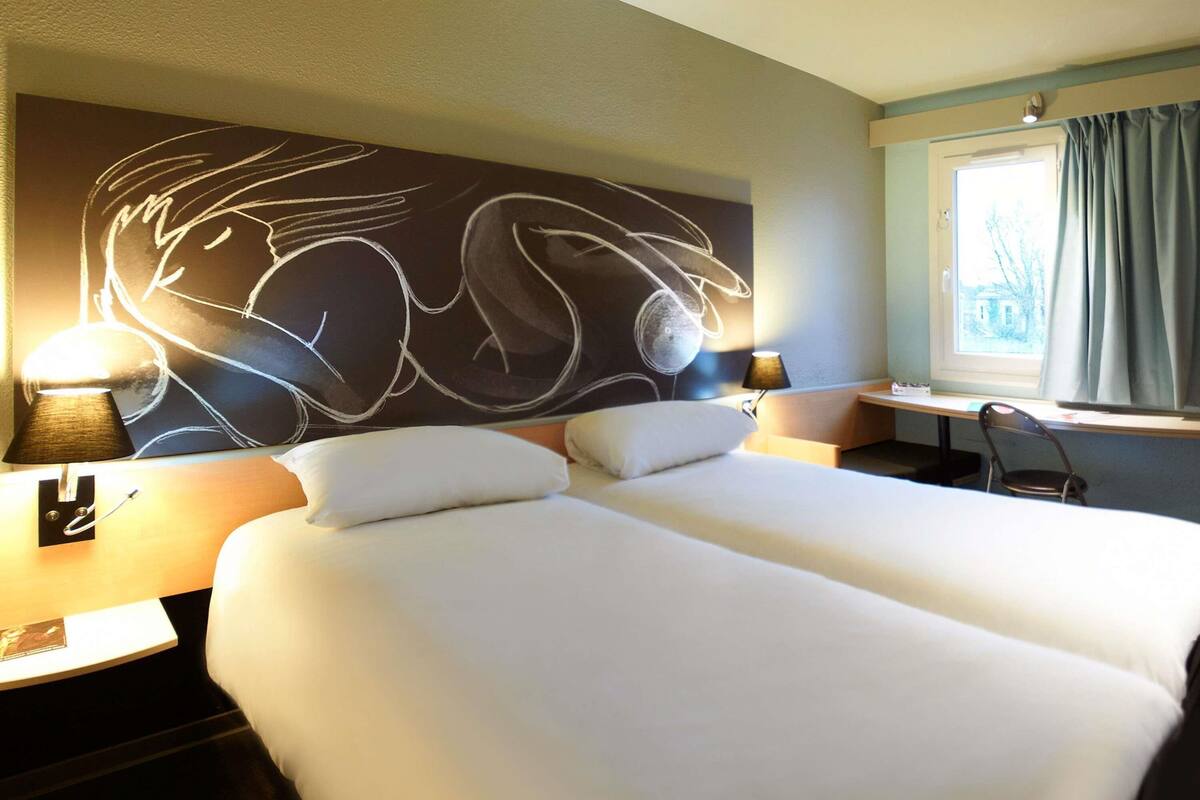 Habitación de hotel con dos camas, un escritorio, una silla, un cuadro abstracto grande en la pared y una ventana con cortinas.