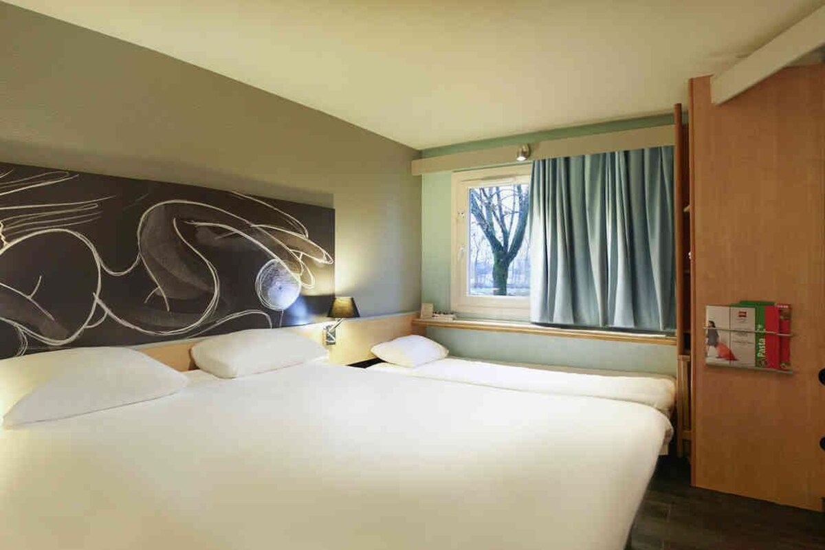 Una habitación de hotel con una cama grande, una ventana con cortinas, una lámpara de pared y una estantería con libros.